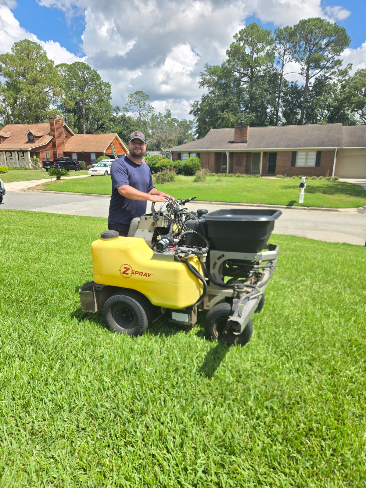 A sunny day scene of a professional applying fertilizer to a large, healthy, green lawn. The image should show the fertilizer being spread evenly and the lawn looking lush and vibrant. Include a sunny sky and clear blue clouds in the background.