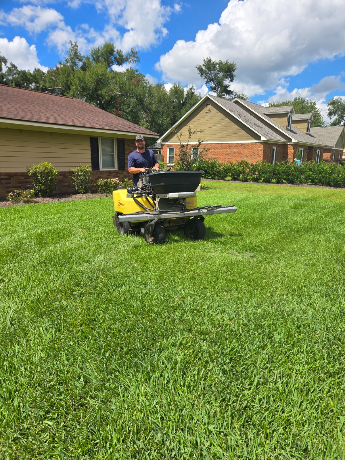 A sunny, outdoor scene with a person applying fertilizer to their lawn using a spreader. The lawn should be bright green and healthy, with a backdrop of a blue sky and fluffy clouds.