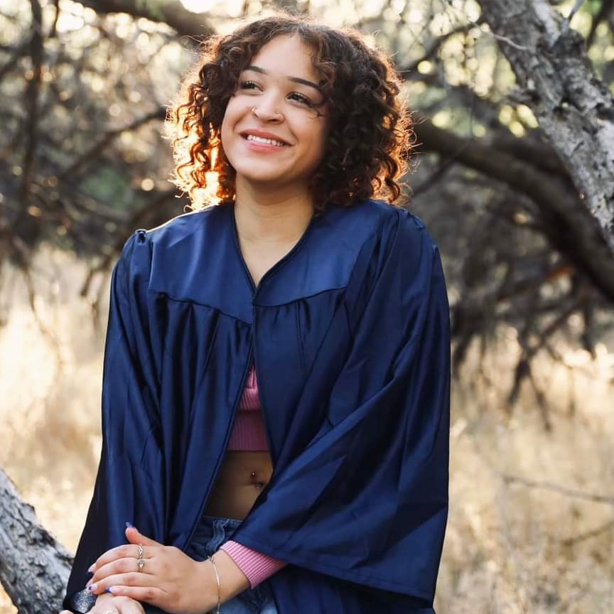 A graduate in a cap and gown stands proudly on a university campus, holding their diploma with a big smile. In the background, excited family members cheer and take photos amidst vibrant campus scenery, showcasing a sense of accomplishment.