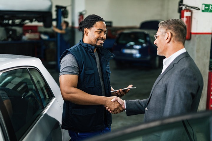 A photorealistic image showcasing a happy customer shaking hands with a mechanic after receiving their repaired vehicle. Both the customer and mechanic should have genuine smiles. The auto repair shop is clean and professional in the background. The lighting is warm and inviting. Color palette focuses on trust and satisfaction. Technical specs: 4K resolution.
