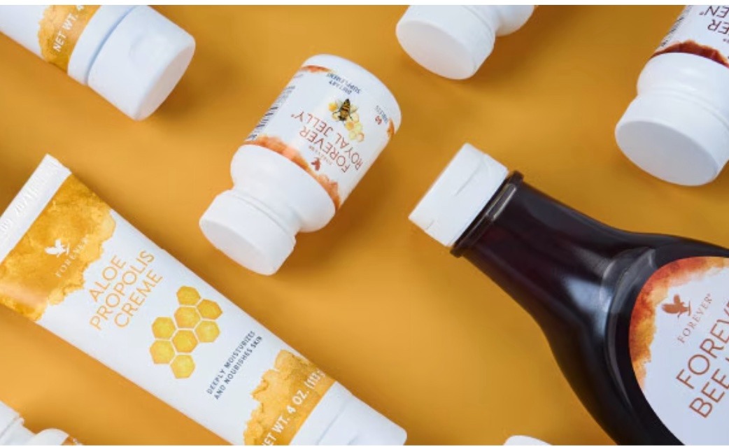 A beautiful photograph of a beehive with golden honeycomb and bees in flight. A jar of Forever's honey and a bottle of bee pollen supplement are placed in front of the hive, showcasing the natural source of these products.