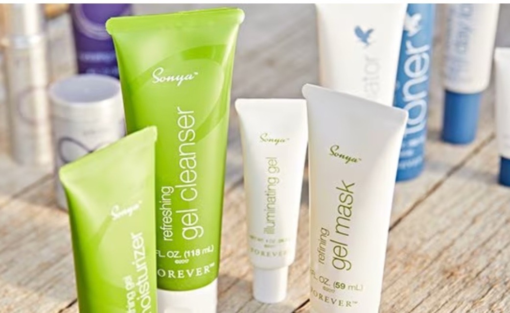 A close-up photo of a selection of Forever's aloe vera products, including soap, shampoo, hand cream, and a bottle of aloe vera juice. The products are arranged on a table with a natural background of green leaves and sunlight.