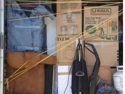 A team of movers in uniform are carefully packing fragile items into boxes while another team member is loading the boxes into a large moving truck. The truck is parked in front of a house, with a sunny, suburban neighborhood in the background.
