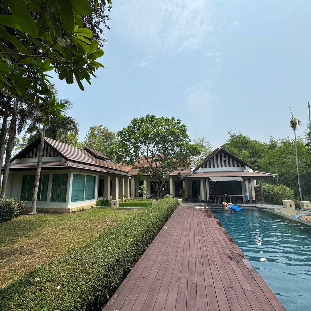 A photorealistic image of a luxurious two-story house with a swimming pool, overlooking the Ping River in Chiang Mai, Thailand. The house should have a modern design with large windows offering stunning river views.  The sky should be clear and sunny, and lush green trees should surround the property. The image should emphasize the tranquility and natural beauty of the location.