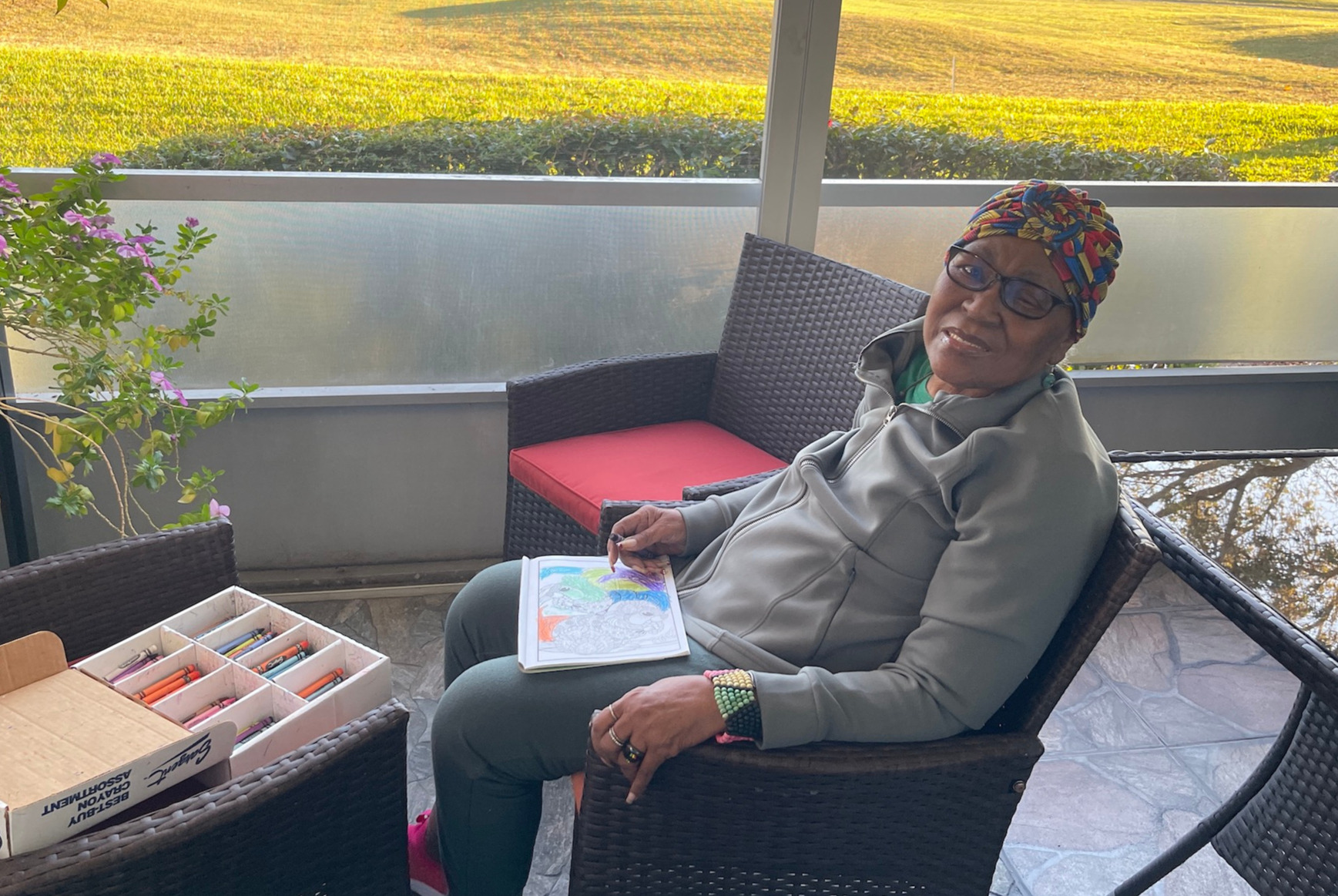 A photorealistic image representing Tina Brown, a certified home health aide, caring for an elderly gentleman in his home. The composition highlights the warmth of their interaction, with Tina smiling and providing support as he reads a book. The lighting is soft, creating an intimate and comforting environment. The color palette consists of warm earth tones with greens and browns, invoking a sense of safety and comfort. The scene includes a cozy living room with a couch, bookshelf, and family memorabilia in the background. The photographer’s angle should be slightly above eye level, focusing on the genuine connection between caregiver and client. The image should be high quality, in 4K resolution.