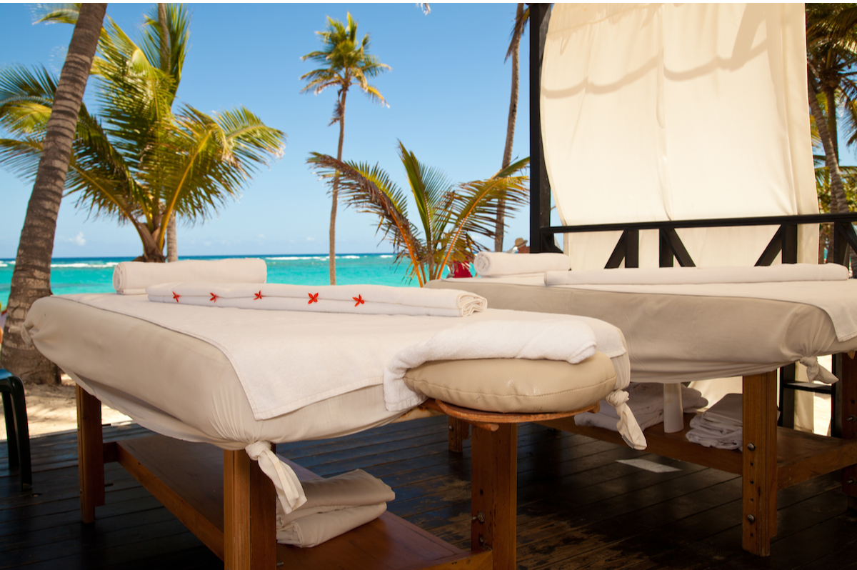 Imagine a beautiful scene representing our mission at Andrea's Therapeutic Massage: a lush beach near the Negrl Tree House with customers joyfully receiving massages. Capture soft, golden sunlight illuminating the space, creating a harmonious vibe filled with tropical plants and serene water in the background. The focus on clients enjoying their treatments will highlight the comfort and tranquility of the services provided. The colors should be vibrant yet soothing, emphasizing relaxation. This high-quality, 4K resolution image should evoke a sense of peace and satisfaction.