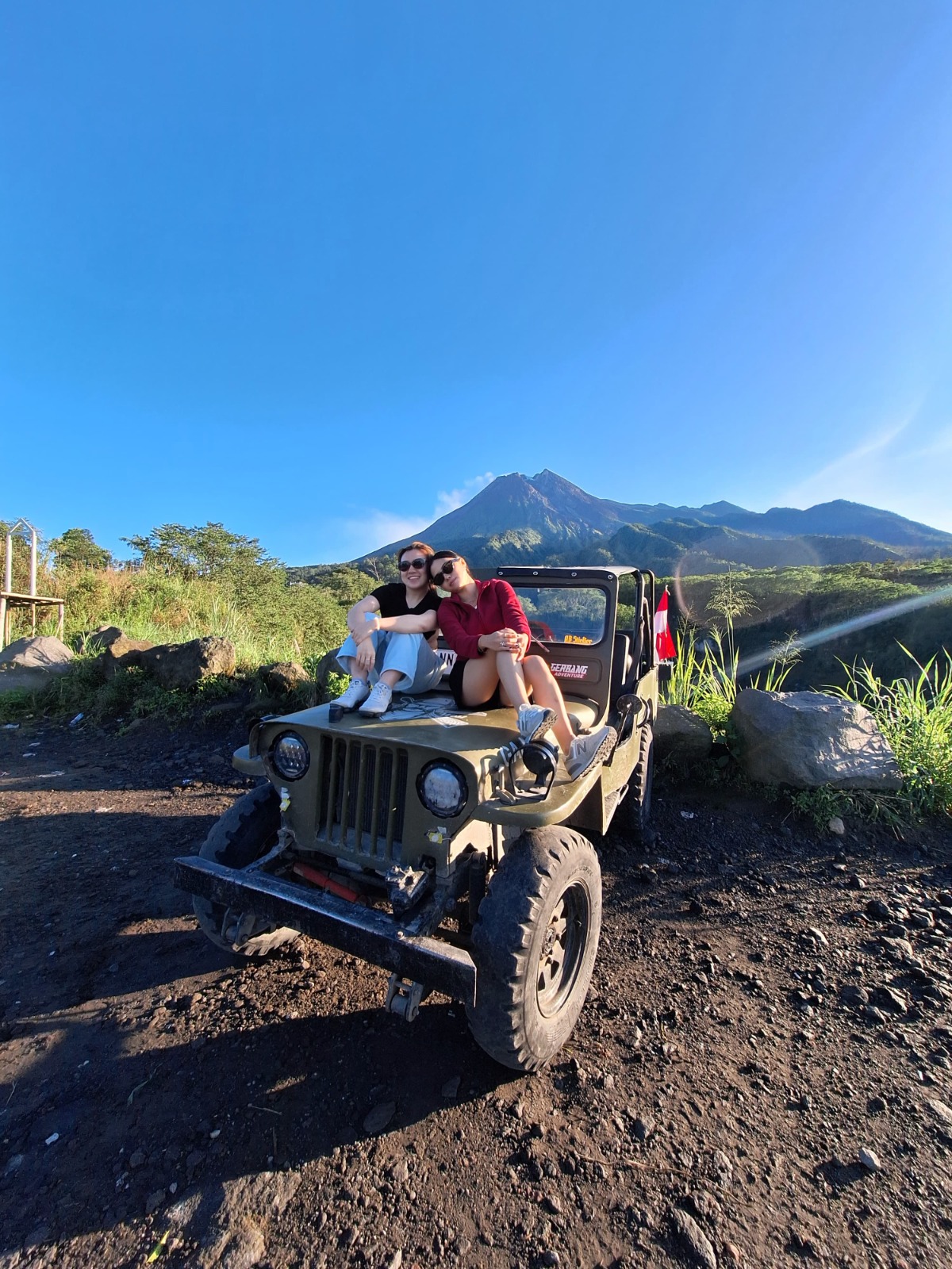 A nostalgic image showing a group of friends reminiscing and laughing after a thrilling Jeep tour at Mount Merapi. The scene captures them with their arms around each other, smiling and joyful against the backdrop of the beautiful volcano. The lighting should be warm and inviting, evoking a sense of camaraderie. Elements like their cameras and adventure gear could be included to signify the journey. Aim for a photorealistic style that evokes emotions and captures the essence of shared experiences.