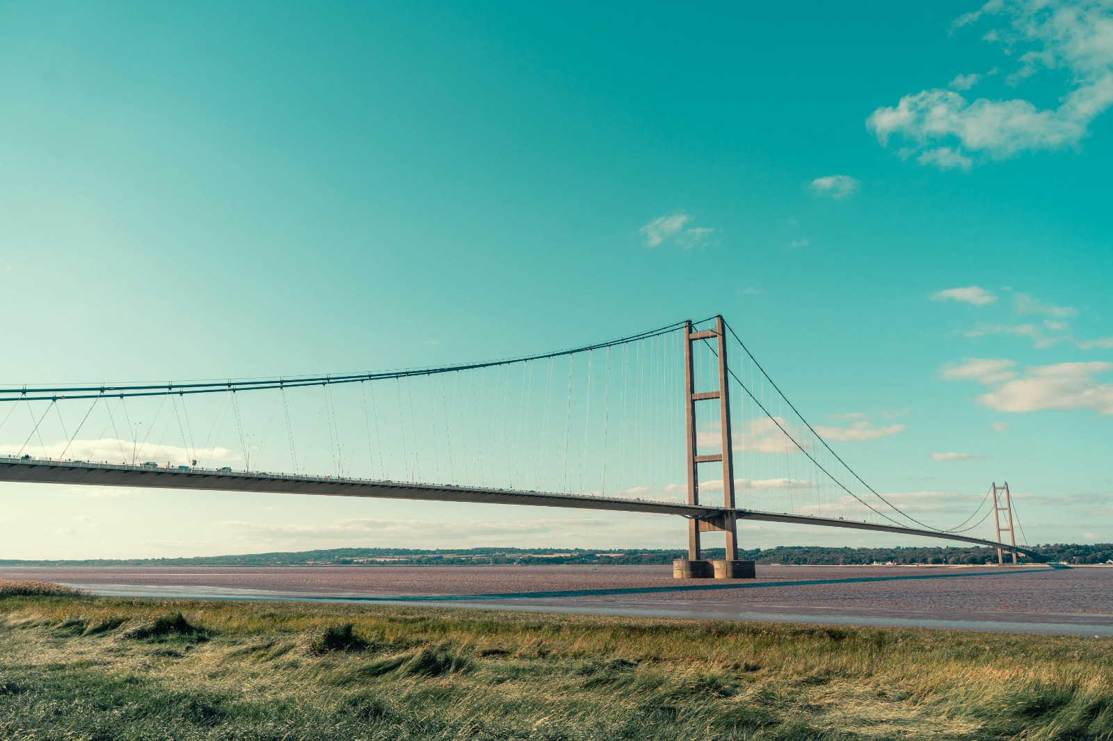 A panoramic view of the Humber Bridge stretching across the River Humber, with lush green landscape on either side. The sky is clear with soft clouds, and people walking across the bridge, capturing the essence of leisure and nature.