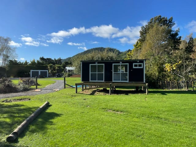 A beautiful, photorealistic image of a cozy sustainable cabin surrounded by nature, emphasizing its organic integration into the environment. The architecture features large windows and a wooden deck, inviting guests to unwind outdoors. The lighting is serene, with soft sunlight filtering through the trees, reflecting a harmonious blend of nature and comfort. Rich greens and earth tones dominate the color palette, creating a welcoming atmosphere. Resolution: 4K, high quality.