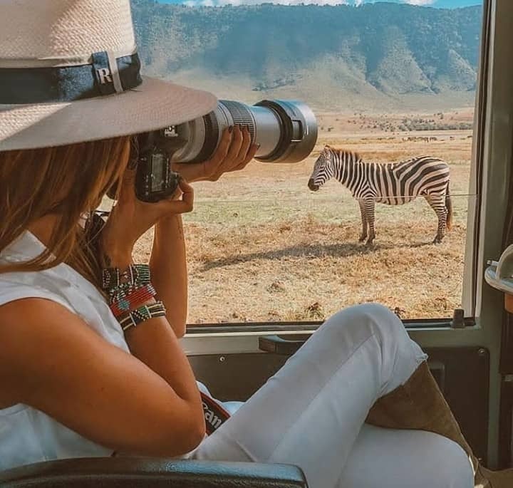 A photographer with professional equipment taking a picture of a lion in the African savanna. The photographer is kneeling down, capturing the lion in its natural habitat. Golden hour lighting and a shallow depth of field. Focus on the beauty of the wildlife and the passion of the photographer. 4K resolution.