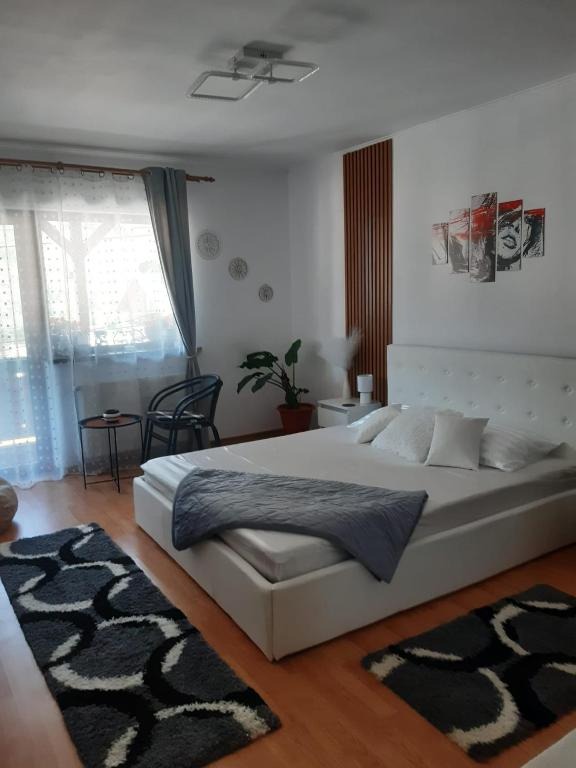 Visualize the interior of one of the bedrooms in "Casa cu 2 Dormitoare La Simo". The scene focuses on a large, comfortable bed with plush pillows and a soft duvet. Sunlight streams through the window, illuminating the room. The color palette is warm and inviting, with light beige and soft blues. A bedside table with a lamp and a book adds a touch of homeliness. Capture the texture of the bedding, the curtains, and the wooden furniture. The lighting should be soft and natural, creating a serene and restful atmosphere. Aim for a photorealistic, high-quality image.