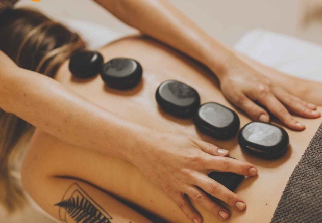  An individual lying face up with warm stones placed along their spine. Eyes closed, expression relaxed, gentle light and soft focus used to evoke a sense of calm. Aromatherapy diffuser nearby releasing gentle mist. 4K resolution.