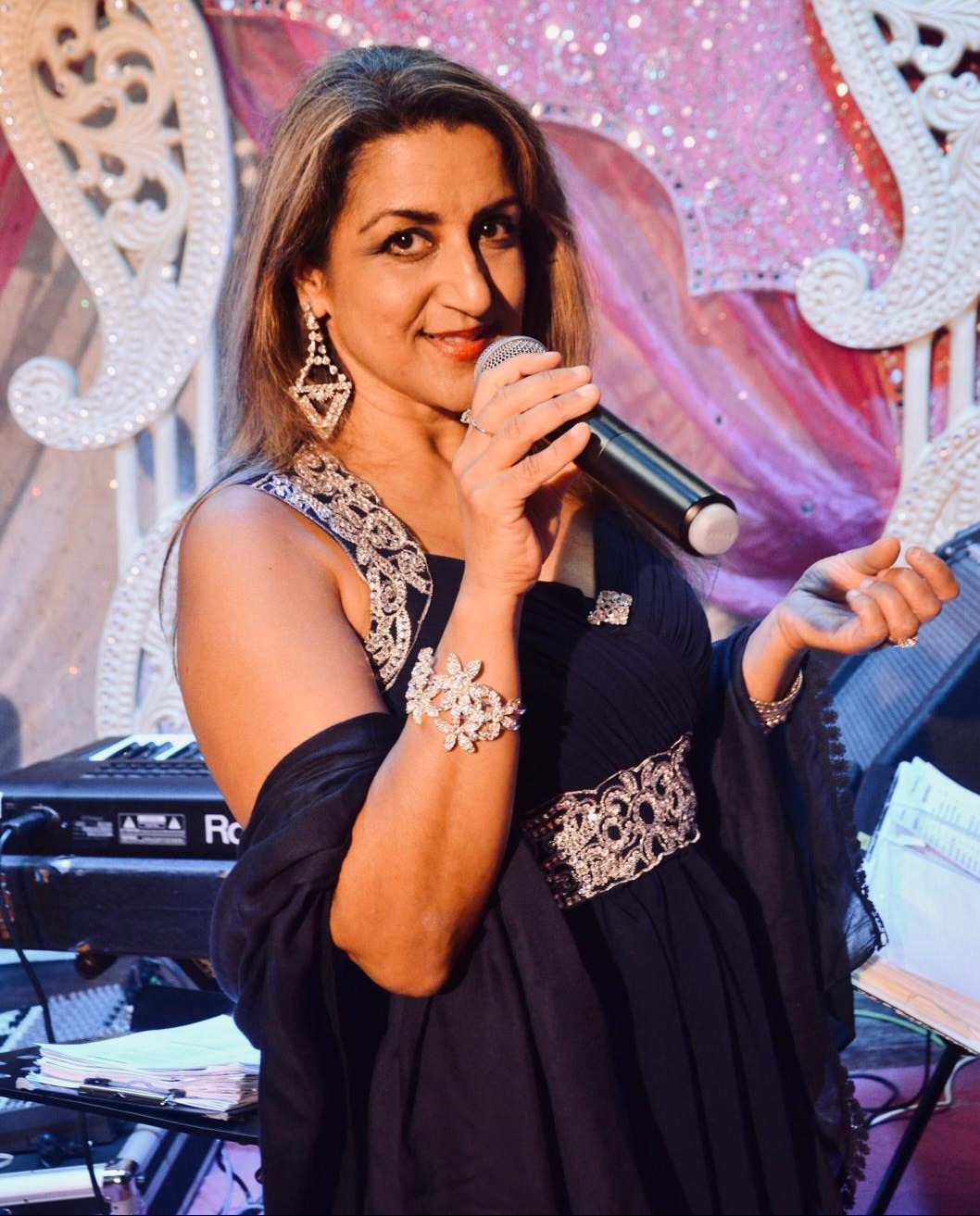A captivating photorealistic 8K hero image showcasing the vibrant energy of Bollywood music. The central figure is Sharan Amelia, a charismatic Bollywood singer in a shimmering, traditional Indian outfit, confidently holding a microphone. Around her, a dynamic live band is performing with enthusiasm, featuring traditional Indian instruments like the sitar and tabla, alongside modern instruments like guitars and drums. The background is a blend of soft-focus stage lights and a cheering crowd, creating a sense of celebration and excitement. The color palette is rich and warm, with golds, reds, and vibrant blues dominating the scene. The camera angle is a medium shot, capturing the singer's captivating presence and the band's energetic performance. Hyperrealistic details should emphasize the texture of the fabrics, the gleam of the instruments, and the expressions of the performers. The overall style is reminiscent of a Bollywood movie poster, conveying joy, festivity, and high-quality entertainment.