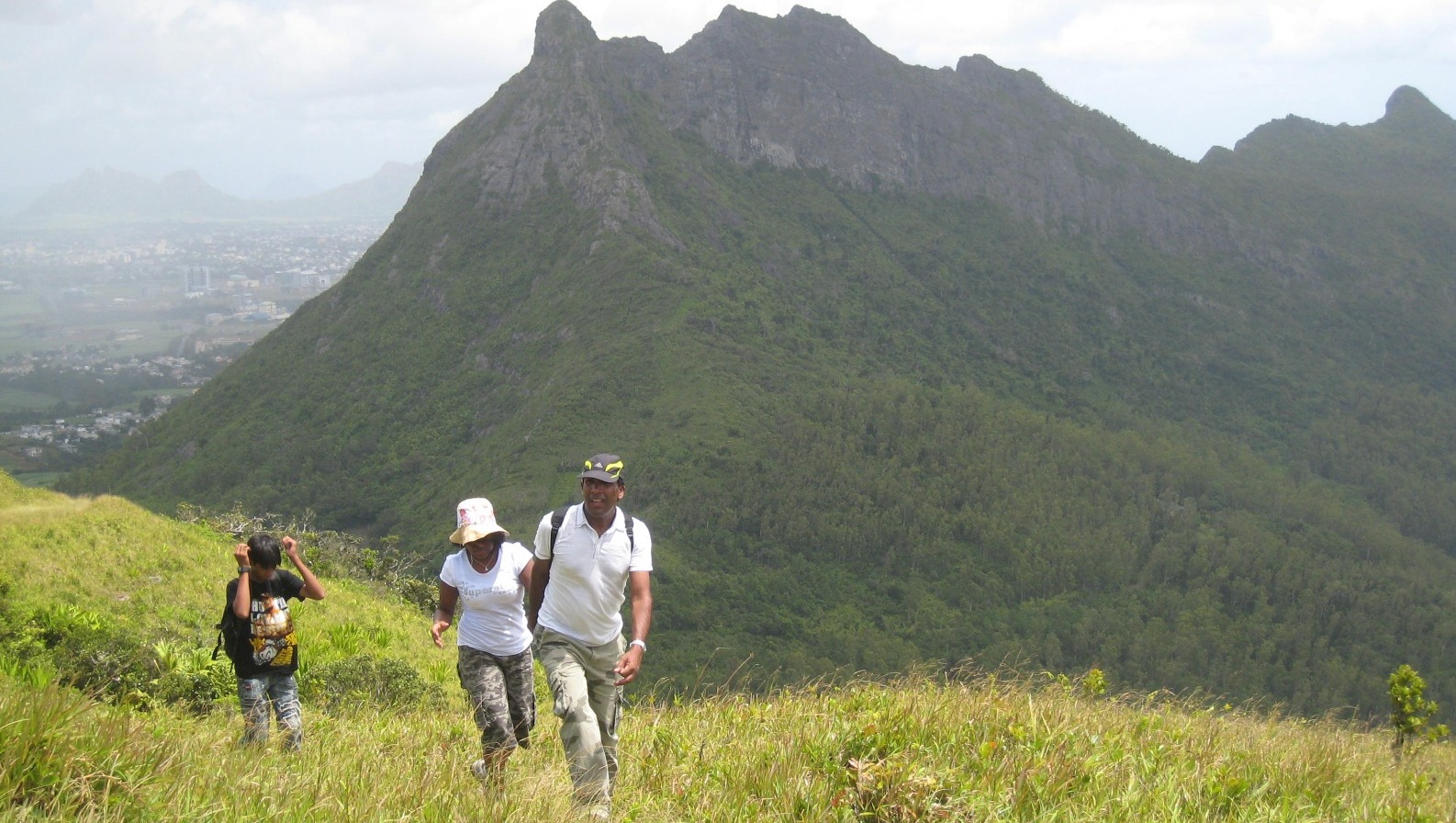 Photorealistic image showcasing a winding hiking trail through a dense Mauritian forest. Focus on the textures of the leaves, trees, and rocks. The lighting should be dappled, with sunlight filtering through the canopy. A small group of hikers is visible in the distance, adding a sense of scale and adventure. 4k resolution.