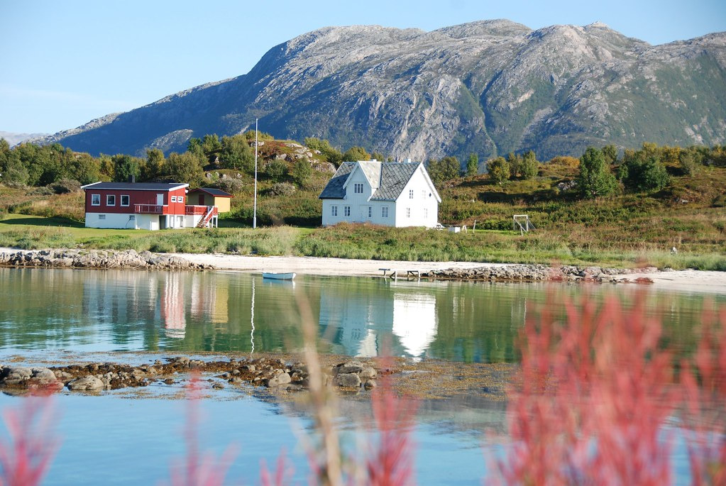 Create an intimate image showcasing traditional Norwegian structures amid the stunning landscapes of Lofoten. The picture should depict a charming red fishing cabin nestled along an emerald coastline under vivid blue skies. Include elements such as fishing nets and traditional boats, with the crisp textures of wood and weathered walls standing out. The golden light of sunset casts long shadows, enhancing the rich colors while providing a sense of warmth and nostalgia. This photorealistic depiction captures the essence of cultural experiences along the journey, rendered in high resolution.