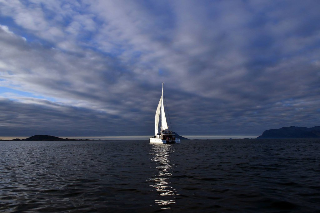 Visualize a sweeping view of fjords and mountains during a cruise from Tromsø to Lofoten. The image should depict lush green mountains reflecting on the still, dark waters of the fjord under a cloudy sky. Soft light filters through the clouds, creating an ethereal ambiance. Include a small fishing boat to emphasize scale, enhancing the sense of tranquility. The scene should evoke feelings of adventure and serenity, maintaining a focus on the natural beauty of Northern Norway. Aim for ultra-high quality to capture intricate details, in 8K resolution.