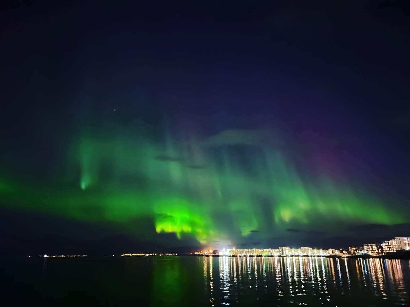 Imagine a serene scene of a group of travelers bundled up in cozy gear, gazing in awe at the auroras dancing across the night sky. The foreground should include a cozy bonfire and hot drinks to suggest warmth amidst the cold. The lighting should highlight the vibrant green, purple, and blue hues of the Northern Lights, contrasting against the dark Arctic sky. The landscape should be snowy and beautifully illuminated by the aurora, creating a magical atmosphere. This photorealistic image should evoke the wonder of this natural phenomenon, making viewers long to experience it themselves.