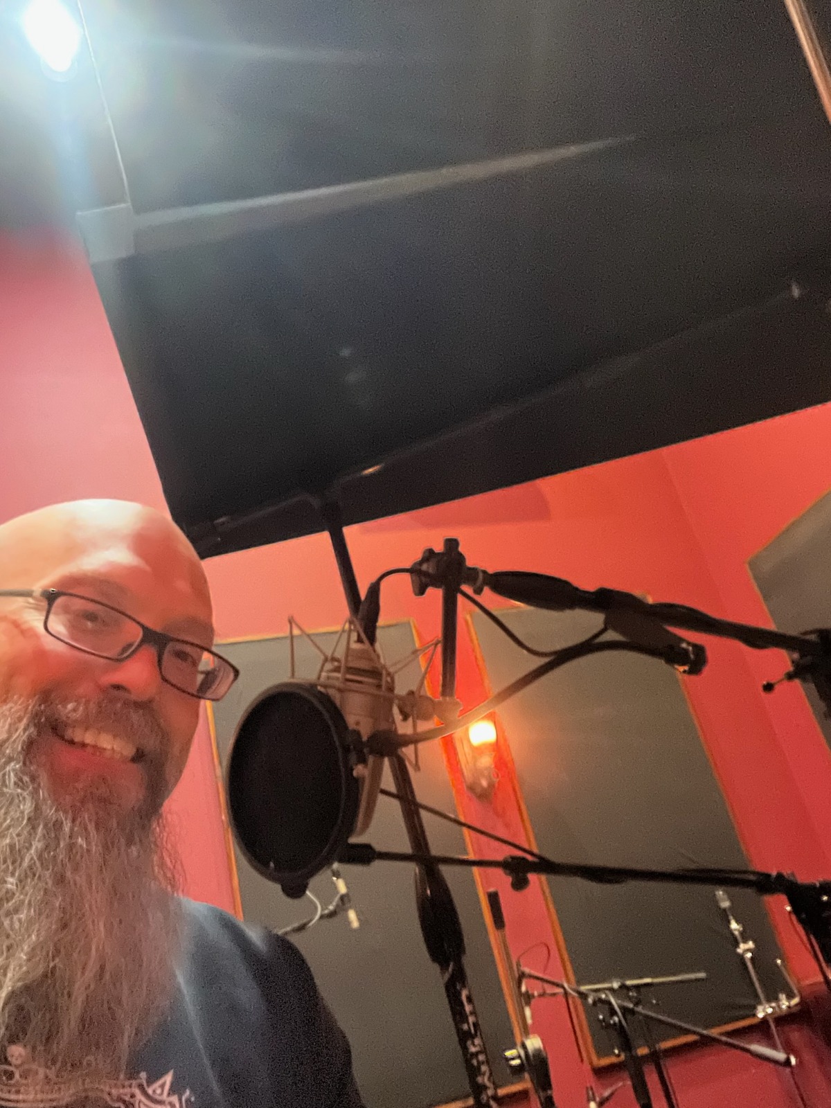 A vibrant and engaging virtual meet and greet scene featuring Frank, the bald musician, sitting in a modern home studio. The lighting is bright and inviting, focusing on Frank's smiling face as he connects with fans through a webcam. The composition includes a computer screen in front of him, displaying excited fans in small video boxes. The color palette is cheerful with pops of bright colors from his T-shirt and the studio decor. Background elements include music awards hanging on the wall and decorative plants adding life to the space. The image should be captured in a close-up perspective to emphasize his relatable expression, rendered in hyperrealistic quality at 8K resolution.