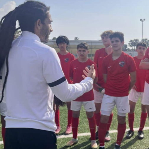 A dedicated football training session with a group of young players practicing skills, led by Auvray, who encourages them. The setting is a lush green field, with a backdrop of mountains, symbolizing growth and development.