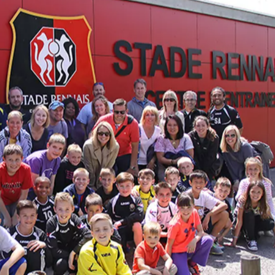 A close-up of a football coach engaged in a dynamic training session with young athletes on a lush green pitch. The composition captures the vibrant energy of the session, highlighting the coach demonstrating dribbling techniques. Soft diffused lighting creates an inspiring atmosphere, enhancing the lush green palette of the field and the bright jerseys of the players. The scene has an uplifting and motivational mood. Captured from a low angle, emphasizing the determination on the coach's face while the players practice in the background. Textures of the grass and the fabric of the jerseys are sharply defined. Surround the scene with trees and goalposts to give a sense of a well-equipped training facility. The style references a mix of sports photography and dynamic portraiture, focusing on sharp detail and color vibrancy. Technical specifications include 8K resolution, hyperrealistic details, and ultra-detailed depiction of the action.
