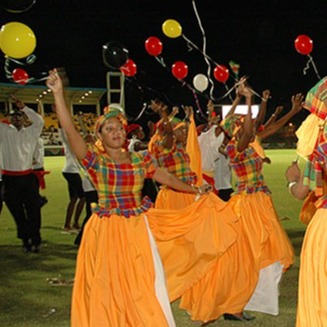 Create a highly detailed, photorealistic image depicting a cultural exchange basketball program in Dominica. The scene should showcase a lively outdoor setting with a basketball court integrated into a tropical environment. Male and female players from U15 to veterans are seen interacting with local residents and participating in traditional Caribbean activities such as dancing and music. The lighting is golden-hour sunlight, creating a warm and inviting atmosphere. The color palette should include vibrant hues like tropical green, ocean blue, and traditional creole clothing patterns. The camera angle is at eye level, capturing the engagement and joy on the faces of the players and local participants. Textures to capture include the court's asphalt, the natural landscape, and the various traditional props like drums and woven baskets. Background elements include lush vegetation, a flowing river, and traditional Caribbean huts. This image should evoke a sense of cultural richness and community, styled in the manner of a National Geographic photograph, with 8K resolution and a hyperrealistic finish.